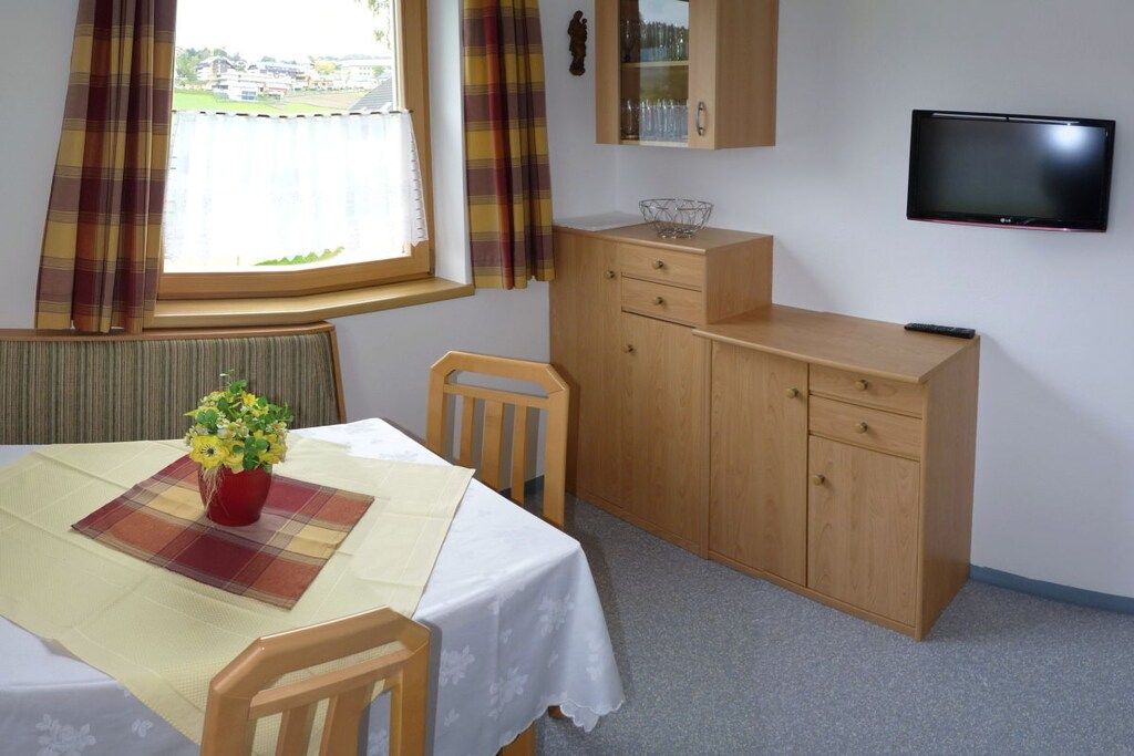 Ein Zimmer mit Esstisch, Stühlen, Fernseher, Schrank und einem fenster mit Vorhängen.