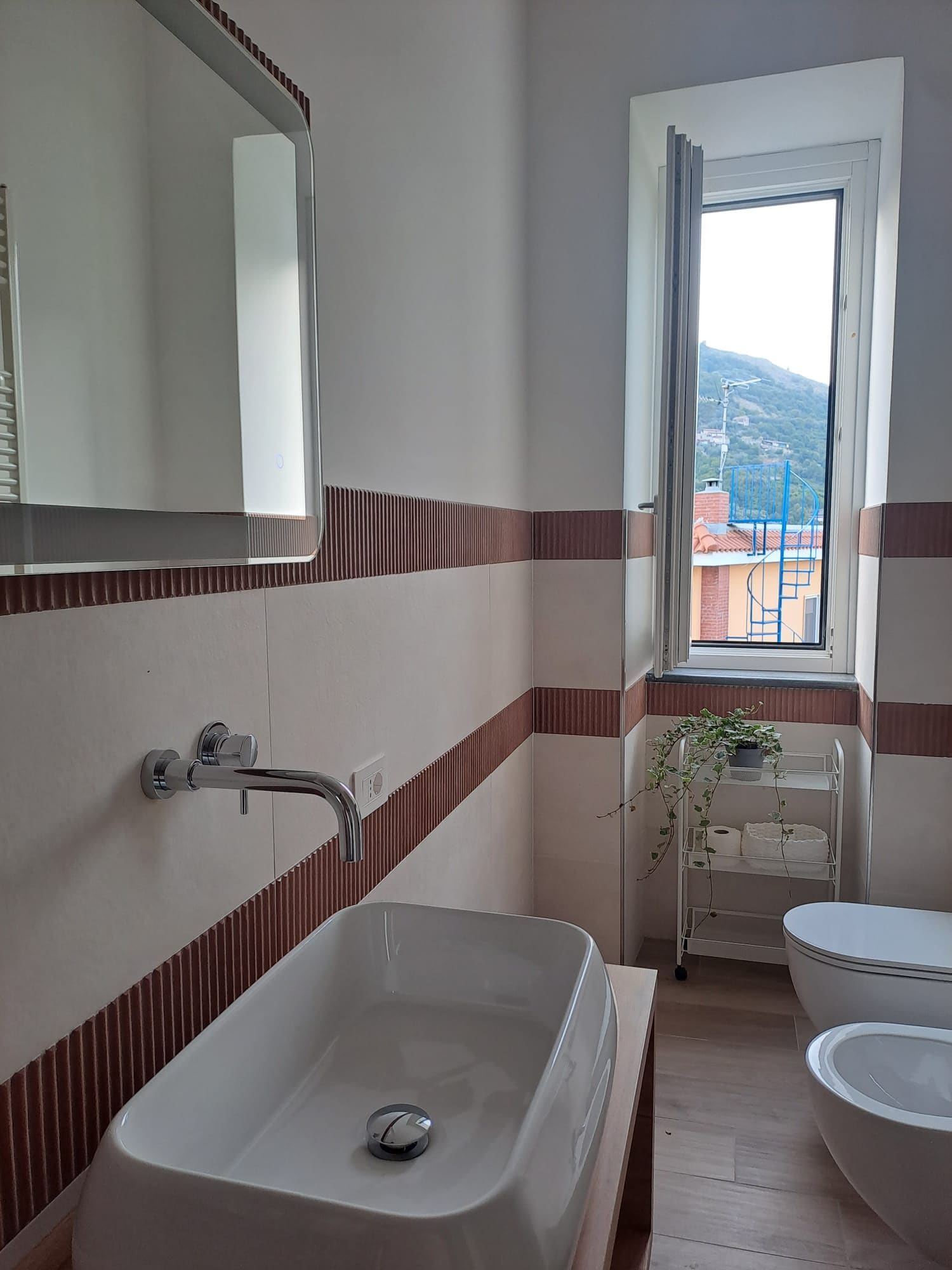 A bathroom with a large sink, a toilet, a mirror, and a window with a view of buildings and hills.