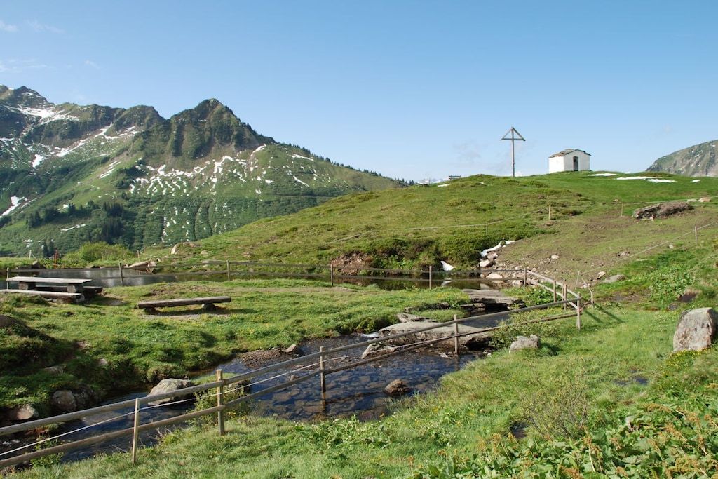 Eine gebirgige Landschaft mit einem kleinen weißen Gebäude, einer Holzbrücke und einem Bach.