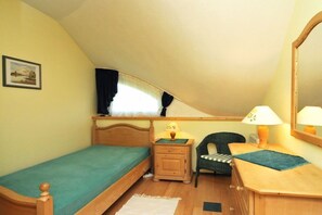Ein kleines Zimmer mit einem Einzelbett, geneigter Decke, einem Nachttisch aus Holz mit Lampe, einem Stuhl und einem Schreibtisch mit Spiegel.