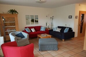 Ein Wohnzimmer mit einem roten Sofa, einem blauen Sofa, einem weißen Sessel und einem grauen Pouf. Es gibt eine Holzvitrine mit Glastüren, eine Stehlampe und ein Bild an der Wand.