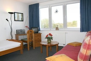 Ein gemütlicher Raum mit einem Bett, einem Schreibtisch mit Radio, einem Sessel, einem Tisch mit Blumen und einem Fenster mit Vorhängen.
