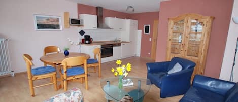 A living room with a glass coffee table, floral sofa, blue armchairs, and a kitchen area with white cabinets and a stove.