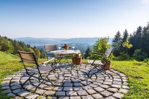 Ein Sitzbereich im Freien mit einem kreisförmigen Tisch aus Steinplatten und vier Stühlen, umgeben von üppiger, grüner Natur und Blick auf ferne Hügel.