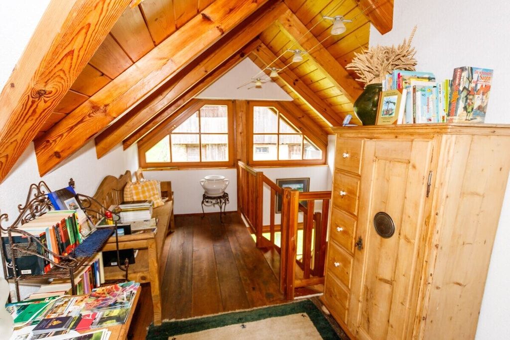 Une pièce mansardée chaleureuse avec des poutres en bois, un lit en mezzanine, une petite table avec des livres et une armoire en bois.