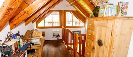 Une pièce mansardée chaleureuse avec des poutres en bois, un lit en mezzanine, une petite table avec des livres et une armoire en bois.