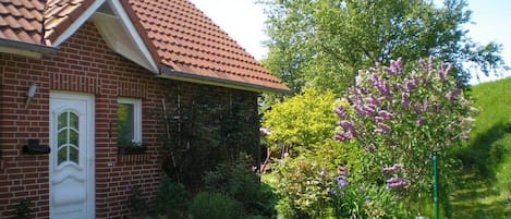Una casa de ladrillo con techo de tejas rojas, una puerta blanca y un jardín con flores moradas.