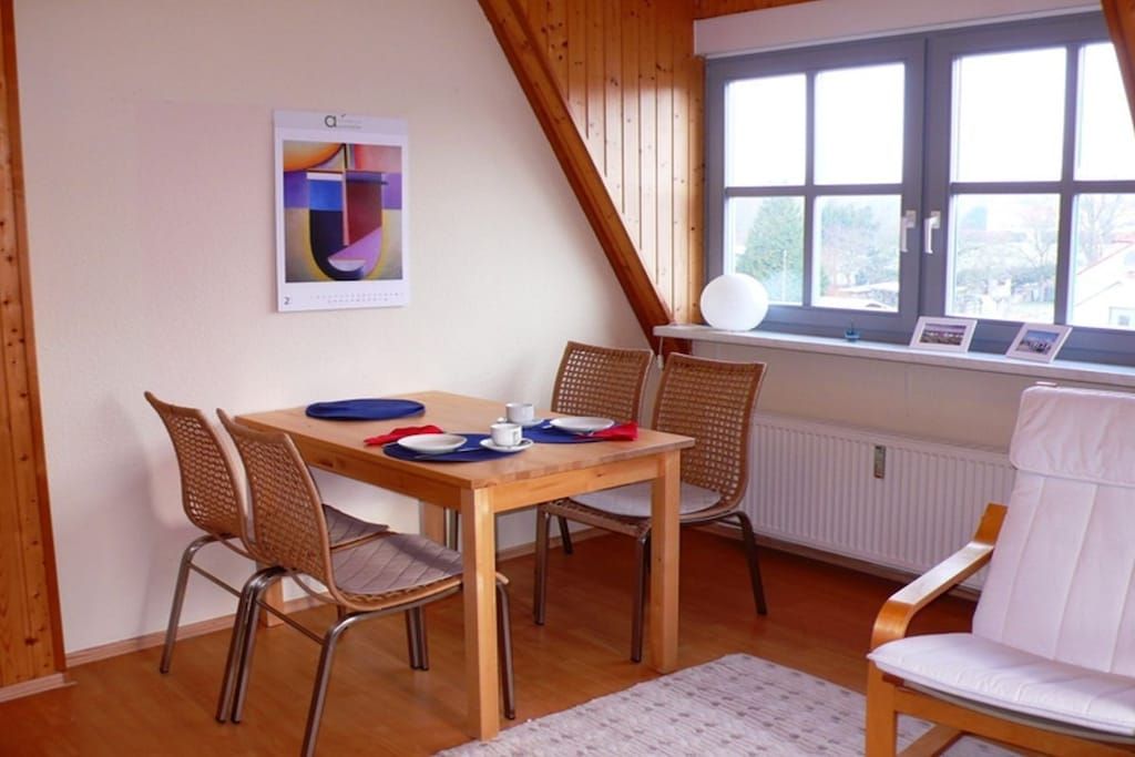 Een eetgedeelte met een houten tafel, rieten stoelen en een witte stoel met kussentje.