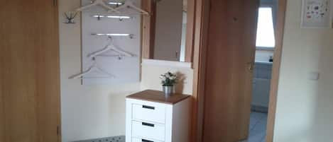 Una habitación con un armario de madera, una mesita de noche blanca con cajones, un perchero y un espejo.
