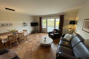 A living room with a sofa, a coffee table, a dining table with chairs, a television, and a window with curtains. - Holiday apartment with 3 bedrooms (Fehmarn)