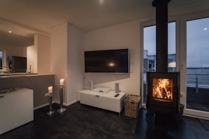 A modern living room with a flat-screen TV, a fireplace, and a large window overlooking a body of water. - Holiday home with 2 bedrooms (Bitterfeld)