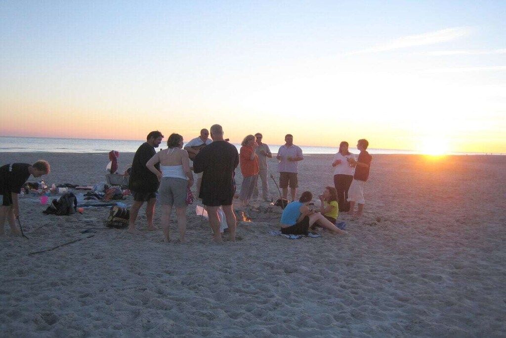 A beach scene with people gathered, some standing and some sitting, with the sun setting in the background.
