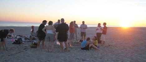 A beach scene with people gathered, some standing and some sitting, with the sun setting in the background.