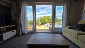 A living room with a sofa, a coffee table, a TV, and a sliding glass door leading to an outdoor area with a pool and greenery. - Killymoon Lodge – Luxury Adults-Only Escape with Private Hot Tub (Newport)
