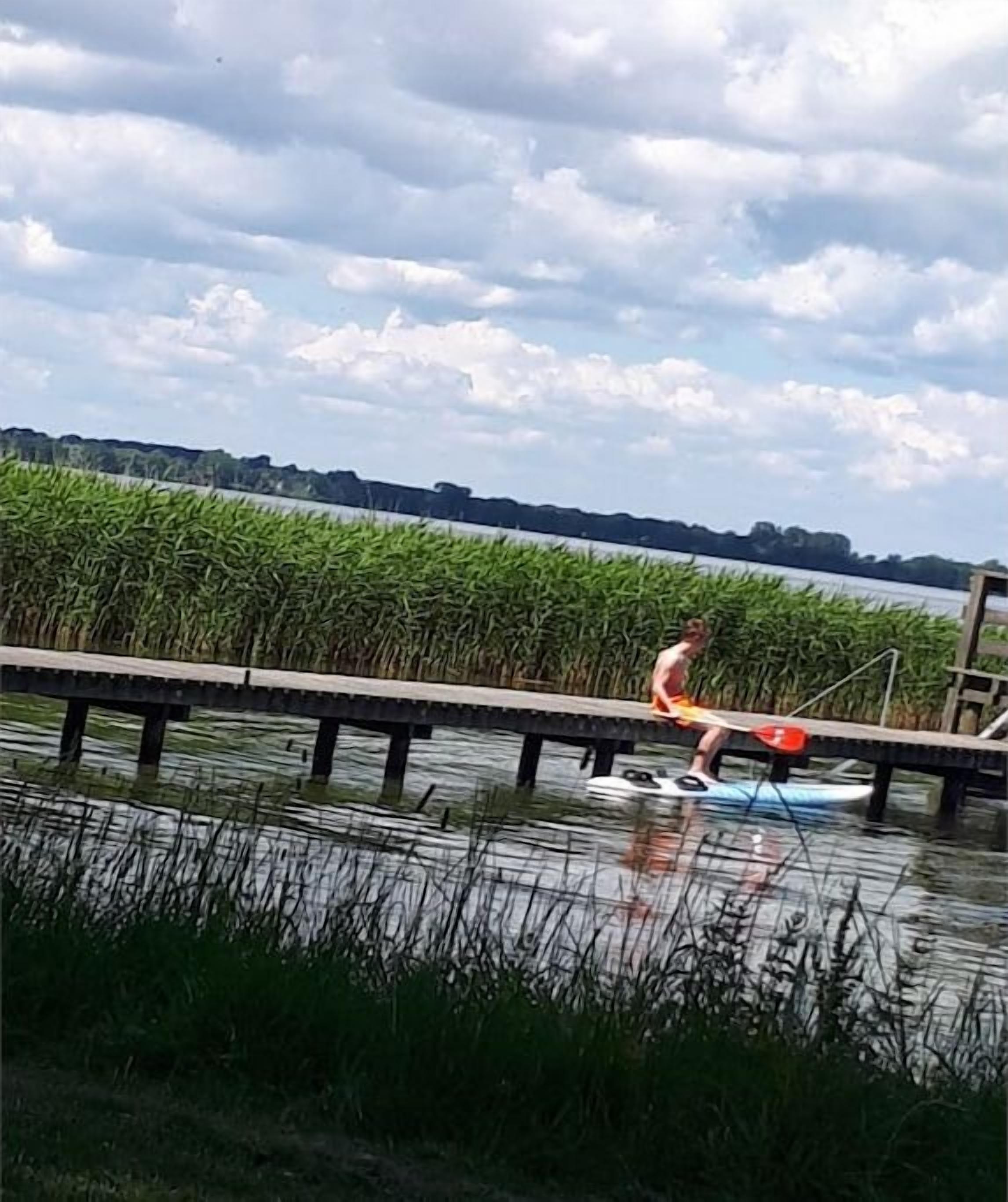 Eine Person steht auf einem Paddleboard auf ruhigem Wasser, im Vordergrund ein Steg und im Hintergrund eine Ufervegetation.