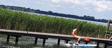 Une personne se tient sur une planche à pagaie, sur une étendue d’eau calme, avec un quai au premier plan et une rangée de végétation en arrière-plan.