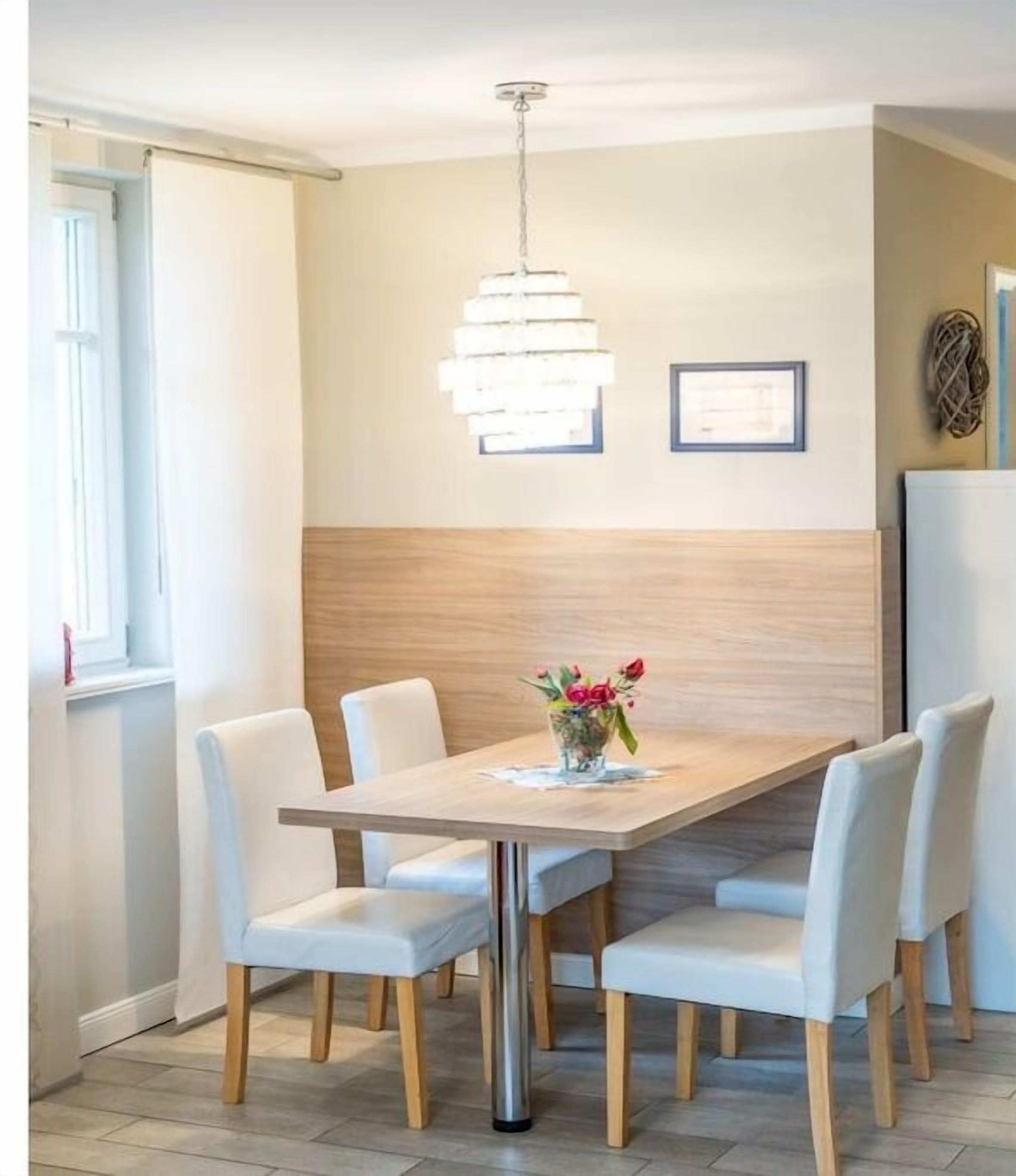 A dining area with a wooden table, white chairs, a hanging light fixture, and a vase with flowers.