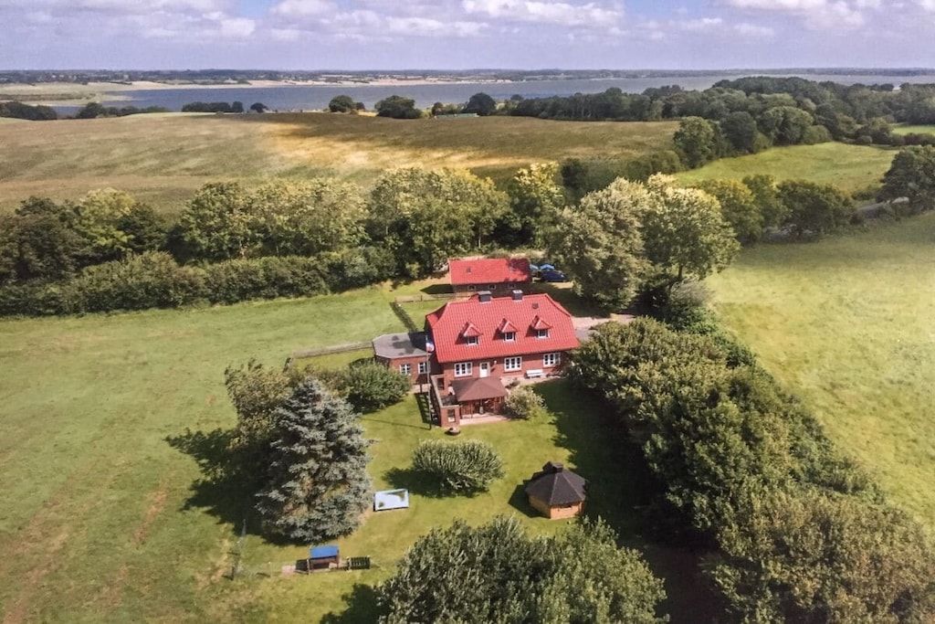 Une grande maison avec un toit rouge, entourée d’un paysage verdoyant et bordée d’une étendue d’eau au loin.