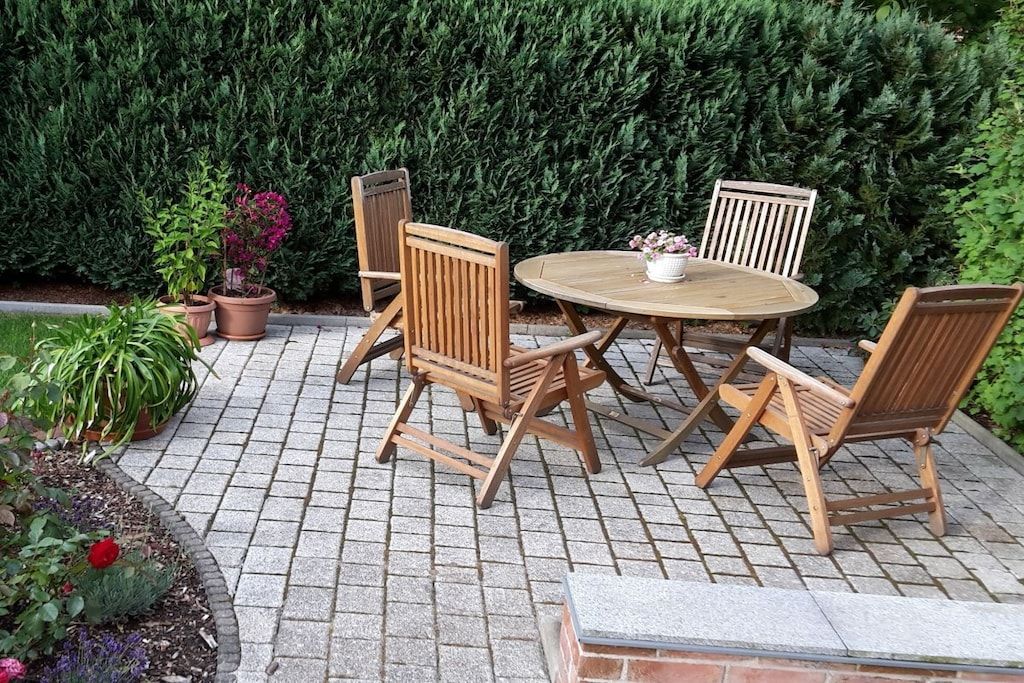 Une terrasse extérieure avec une table ronde et quatre chaises en bois, entourée de plantes en pot et d’une haie.
