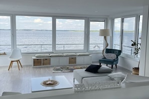 Ein modernes Wohnzimmer mit großen Fenstern, einer Couch, einem Sessel, einer Lampe und Blick auf das Wasser.