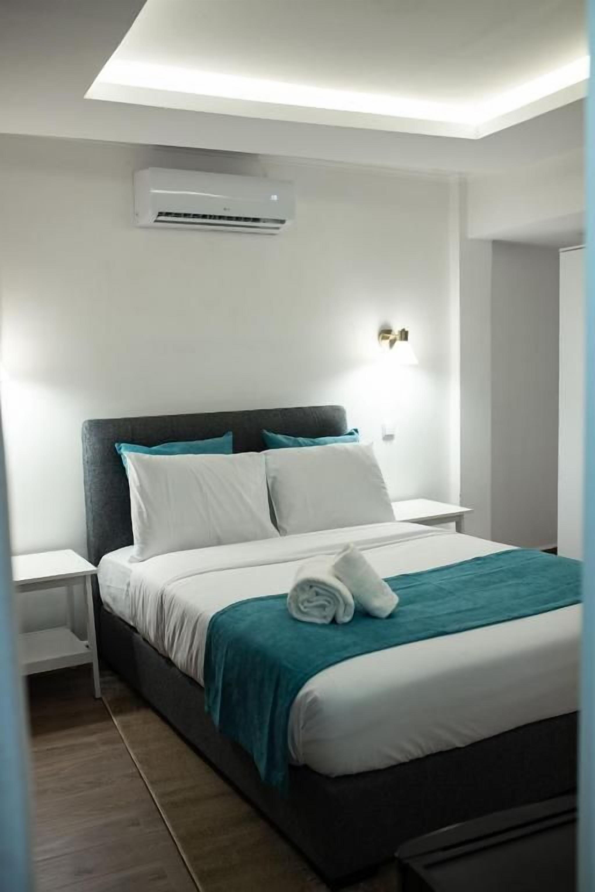 A modern hotel room with a bed, nightstands, an air conditioning unit, and a wall-mounted light fixture.