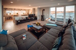 A modern living room with a large sofa, a coffee table, and a television. There is a kitchen area in the background with a large window overlooking a body of water. - Holiday home with 2 bedrooms (Bitterfeld)