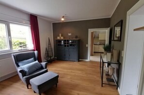 A living room with a grey sofa, a wooden floor, a cabinet with glass doors, and a view to the kitchen. - Holiday apartment with 1 bedroom (Ostseebad Boltenhagen - Tarnewitz)