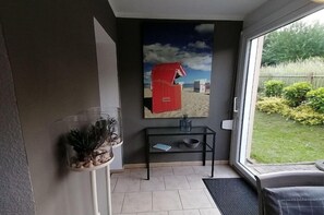 Ein Zimmer mit Glastür, die zu einem Garten führt, ein kleiner schwarzer Tisch mit einer Vase und ein gerahmtes Bild einer roten Strandhütte.