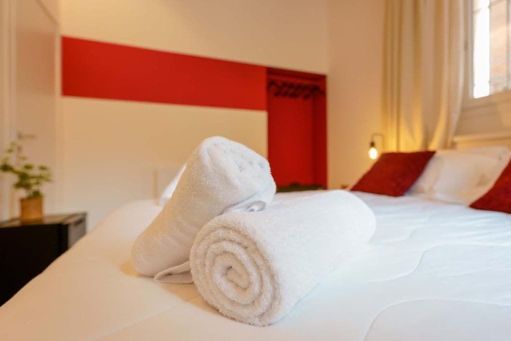 Un lit bien fait, avec des serviettes blanches pliées en rouleaux, un mur d’accent rouge et une lampe de chevet.