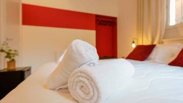 Un lit bien fait, avec des serviettes blanches pliées en rouleaux, un mur d’accent rouge et une lampe de chevet.