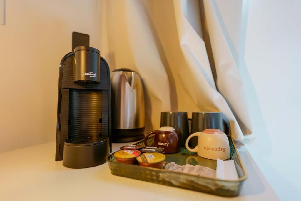 Una cafetera, una olla de acero inoxidable, una taza y una bandeja con recipientes sobre una mesa.