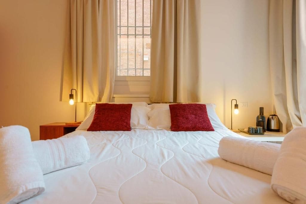 Une chambre d’hôtel avec un lit, deux oreillers, une table de chevet, une lampe et une fenêtre avec des rideaux.