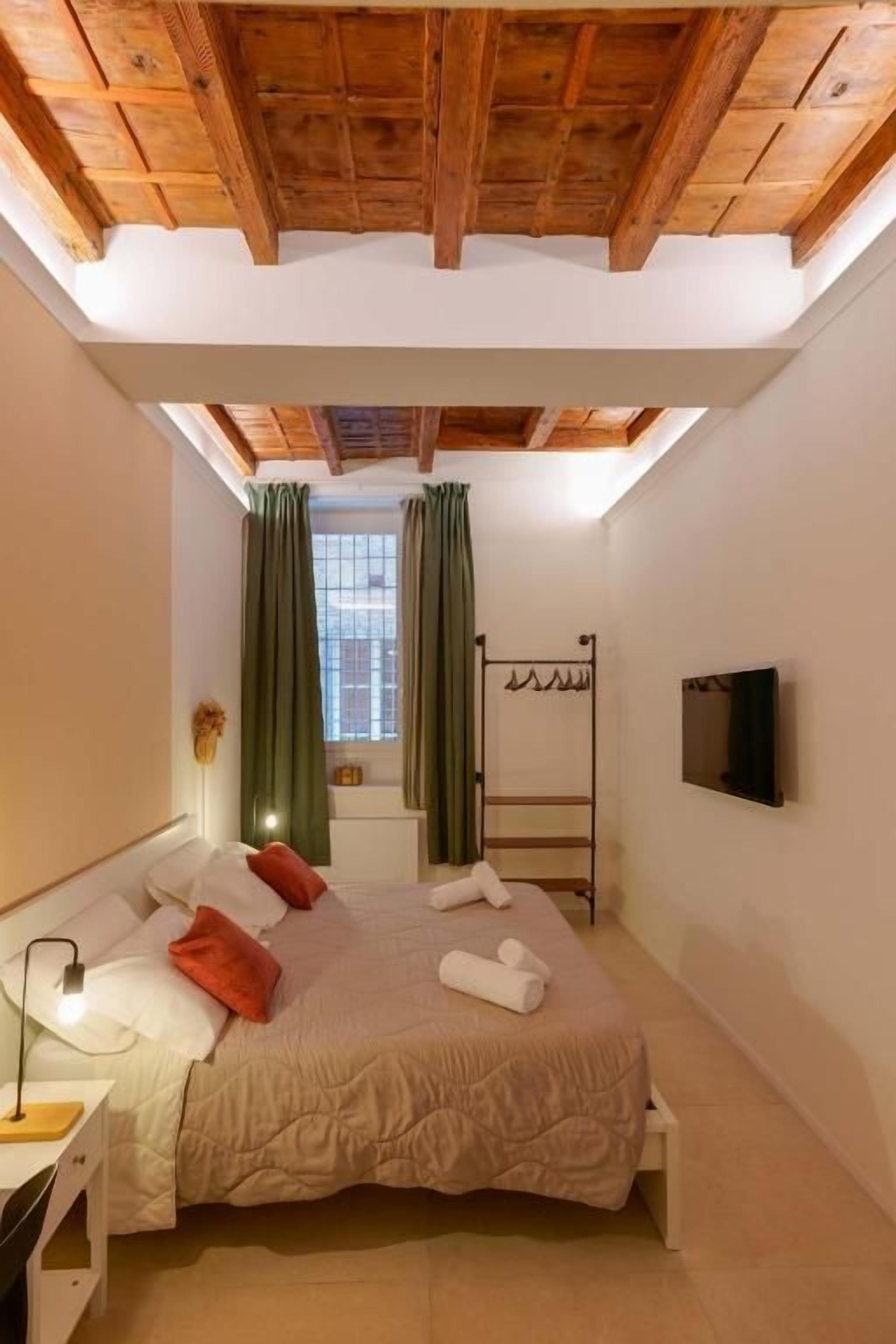 Un dormitorio con cama, mesita de noche, televisor de pared y ventana con cortinas.