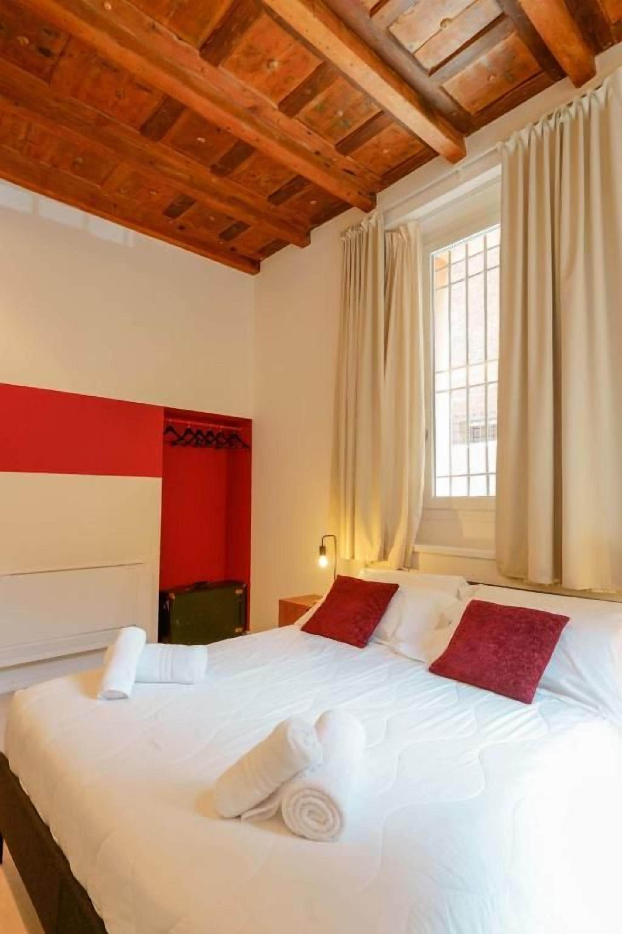 Habitación de hotel con techo de madera, cama con ropa blanca y almohadas rojas, ventana con cortinas color beige y una pared de acento roja.