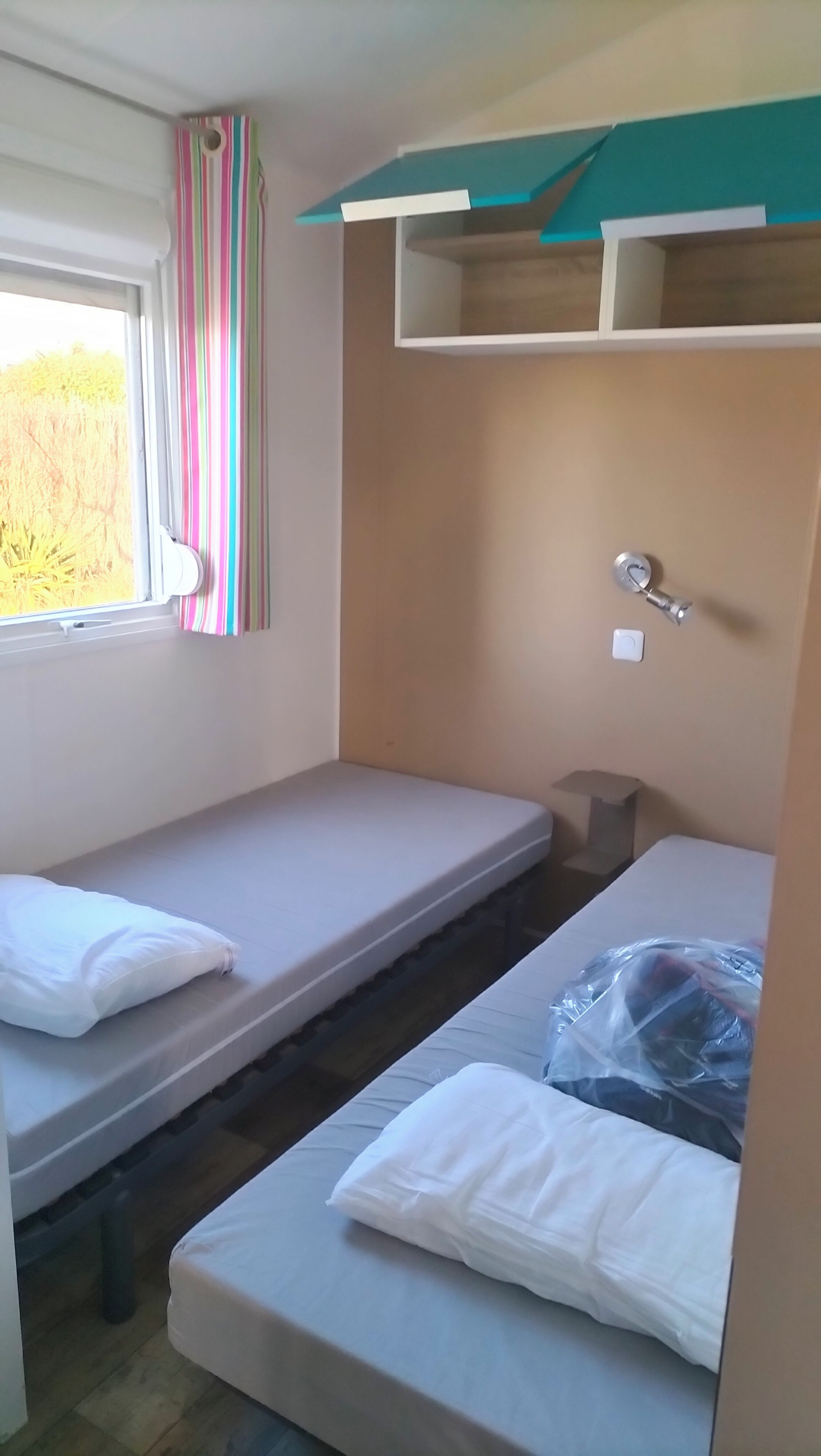 Een kamer met twee eenpersoonsbedden, een raam met gordijnen, een plank boven de bedden en een klein nachtkastje.