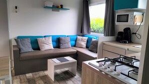 A compact living space with a sofa, a small table, a kitchenette with a stove, and a microwave. - Rent a mobile home (Saint-Hilaire-de-Riez)