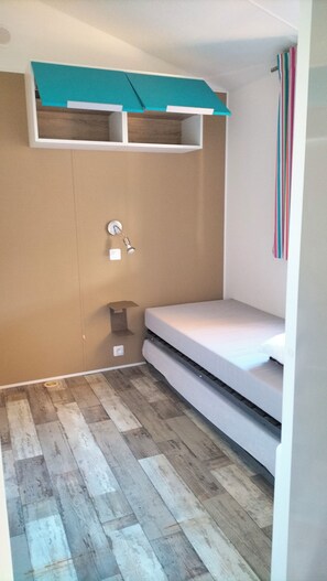 A small room with a bunk bed, a wall-mounted shelf, a light switch, and a striped curtain. - Rent a mobile home (Saint-Hilaire-de-Riez)