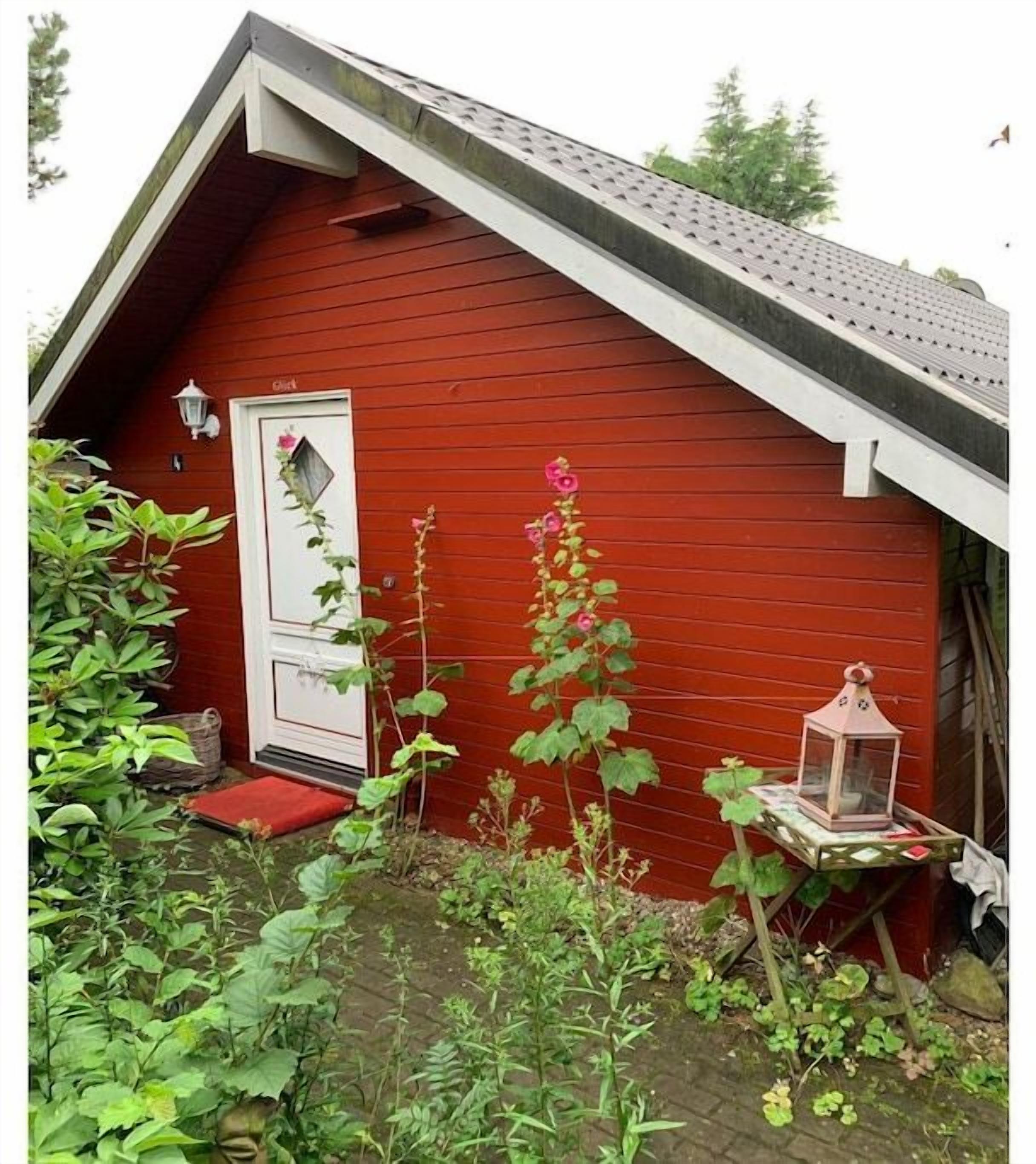 En röd träbyggnad med en vit dörr, en liten trädgård med växter och en lykta på ett bord.