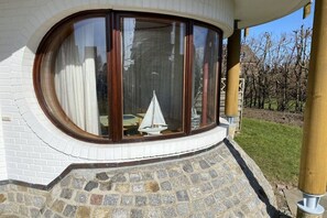 A corner of a building with a curved window, a stone-paved area, and a view of a sailboat inside. - Holiday apartment with 2 bedrooms (Friedrichskoog-Spitze)