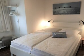 A hotel room with a bed, a bedside table, a lamp, a painting on the wall, and a clothing rack. - Holiday apartment with 2 bedrooms (Friedrichskoog-Spitze)