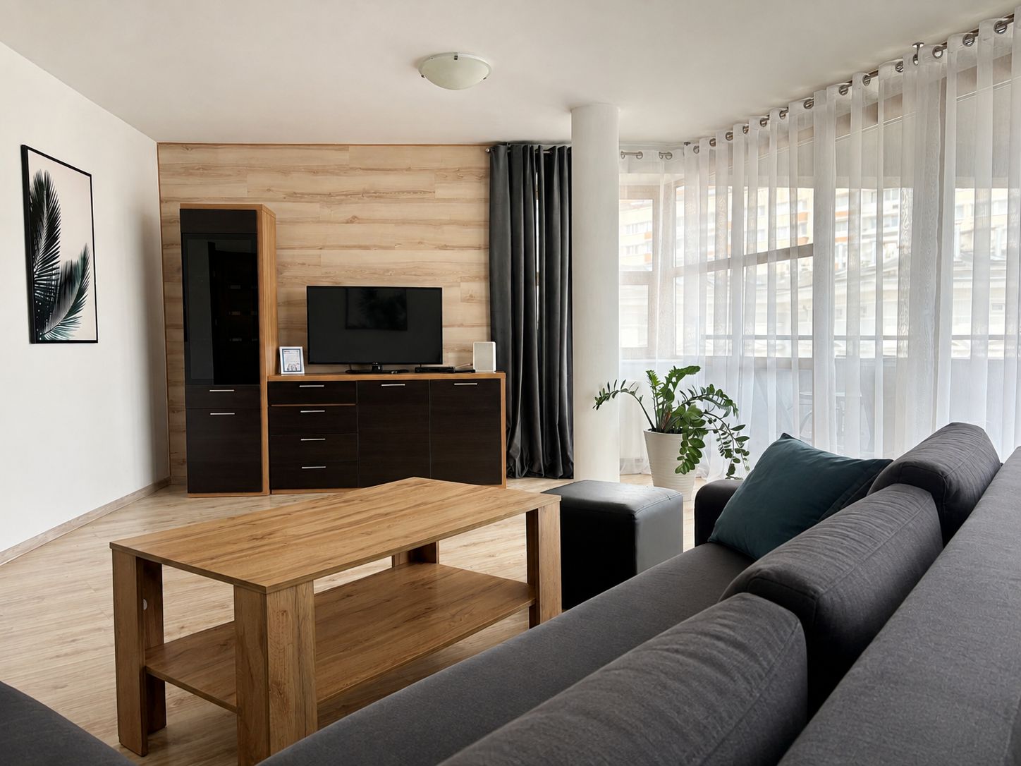 Un salon moderne avec une table basse en bois, un canapé foncé, un téléviseur posé sur un meuble en bois et de fines rideaux sur une grande fenêtre.