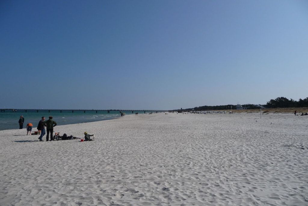 Una amplia playa de arena con cielo despejado, un muelle a lo lejos y algunas personas dispersas a lo largo de la orilla.
