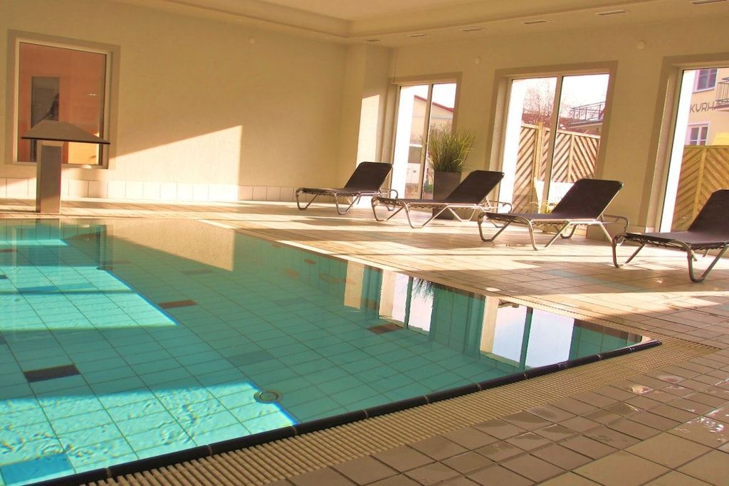An indoor swimming pool with clear water, tiled floor, and sunlit room with windows and lounge chairs.