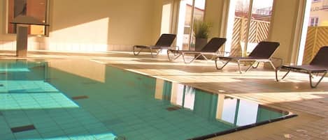 An indoor swimming pool with clear water, tiled floor, and sunlit room with windows and lounge chairs.