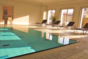 Une piscine intérieure à l’eau claire, dotée d’un sol carrelé et d’une pièce baignée de lumière naturelle grâce à de grandes fenêtres, ainsi que de chaises longues.