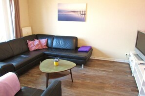 A living room with a sectional sofa, a small round table with a plant, and a flat-screen TV on a stand. - Holiday apartment in Rerik with swimming pool (Rerik)