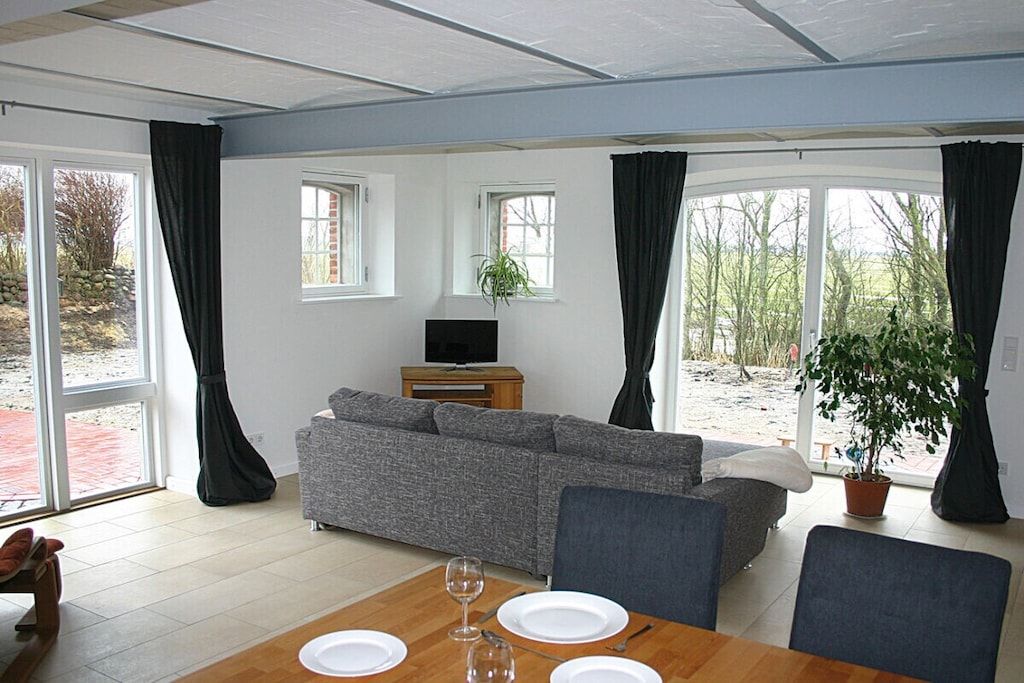 En stue med sofa, spisebord med tallerkener og glas, fjernsyn og store vinduer med gardiner.