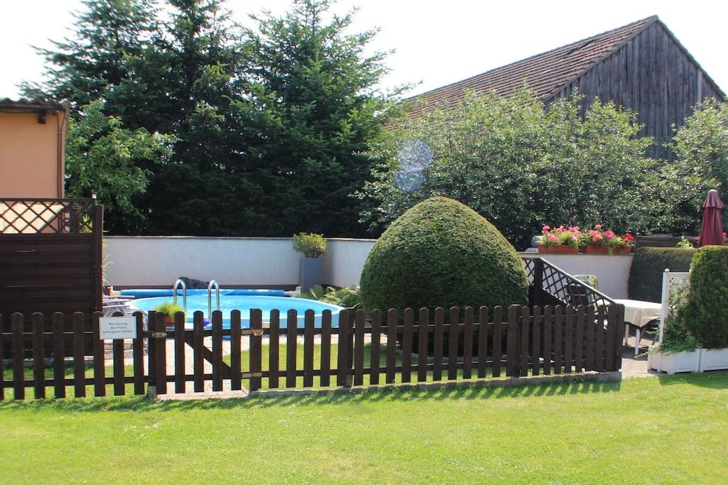 A fenced backyard with a swimming pool, a wooden deck, and a large barn-like structure in the background.