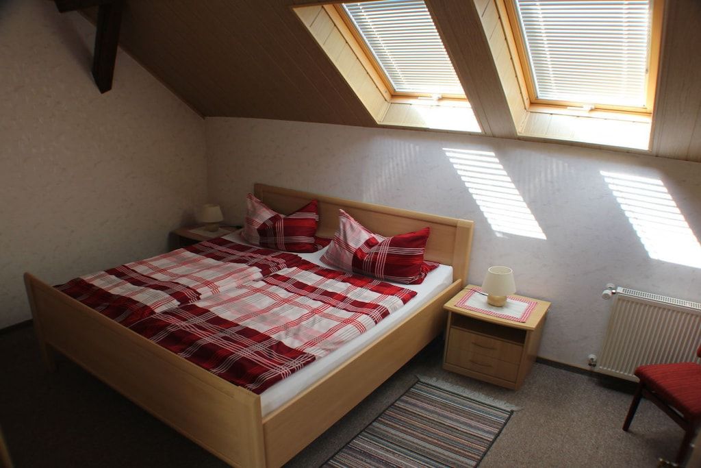 A bedroom with a sloped ceiling, a bed with a checkered bedspread, two bedside tables with lamps, and a radiator.
