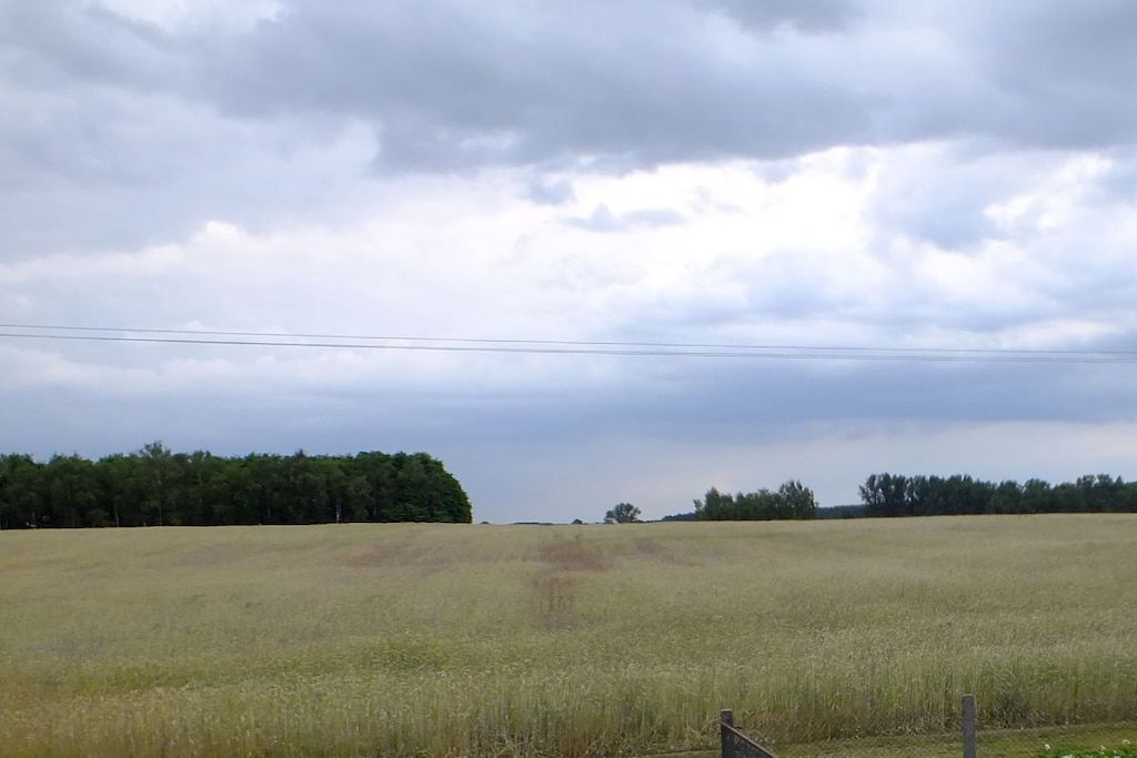 Een uitgestrekt veld met een houten omheining op de voorgrond, onder een bewolkte lucht.